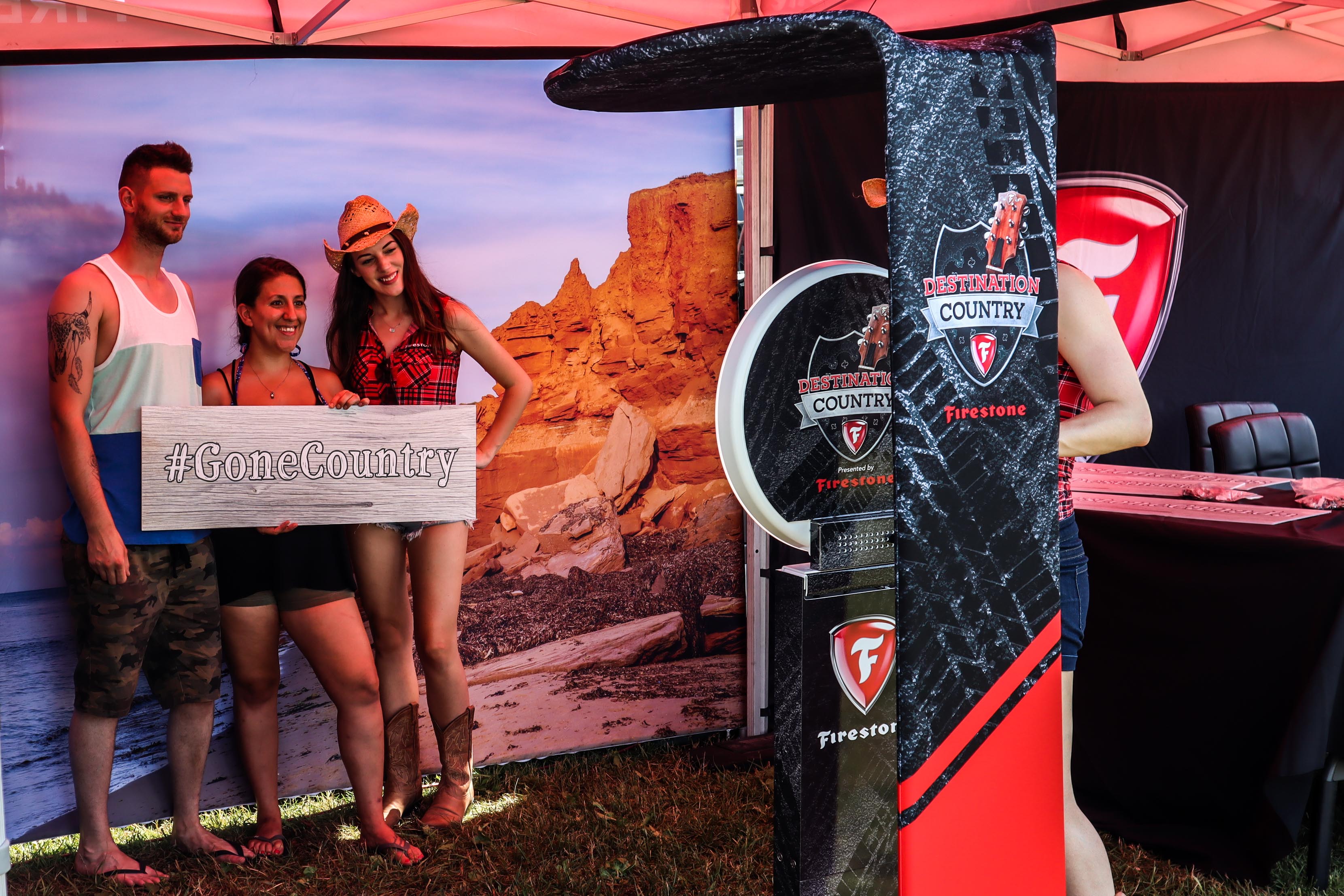 Firestone Destination Country branded photo booth at Cavendish Beach Music Festival — three festival-goers, one in a cowgirl hat, holding a #GoneCountry sign in front of a PEI coastal backdrop